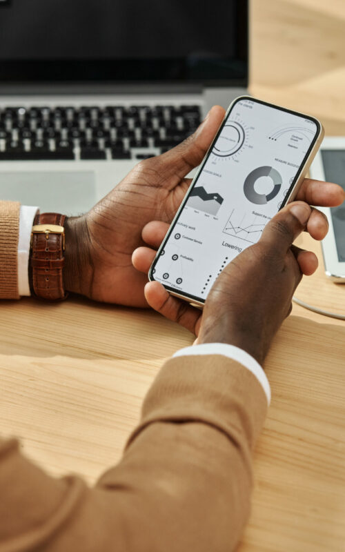 Close-up of African American businessman using smartphone to work with financial graphs while sitting at workplace with computer device